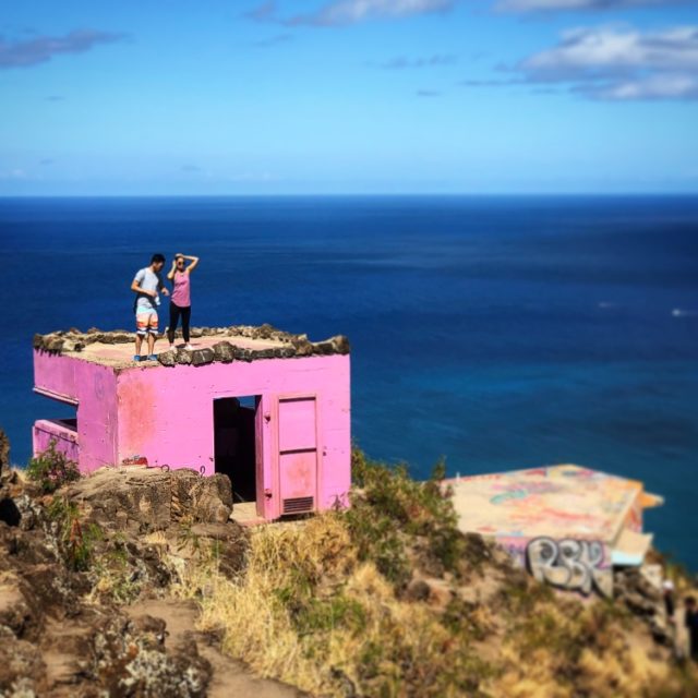 The pink Maili pillbox, or abandoned cement military bunker, is found at the end of a one-mile one-way trail just ten minutes from Ko Olina. In this image, two people are standing on top of the bright pink pillbox with the unending Pacific Ocean behind them.