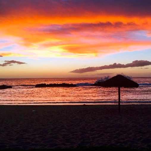 Beautiful Ko Olina sunset with orange and pink clouds, as well as an thatched umbrella silhouette in the foreground.