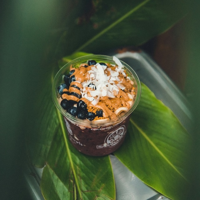 Acai bowl topped with peanut butter, blueberries, and coconut shreds in plastic Sambazon-labeled cup, peaking through bright green leaves